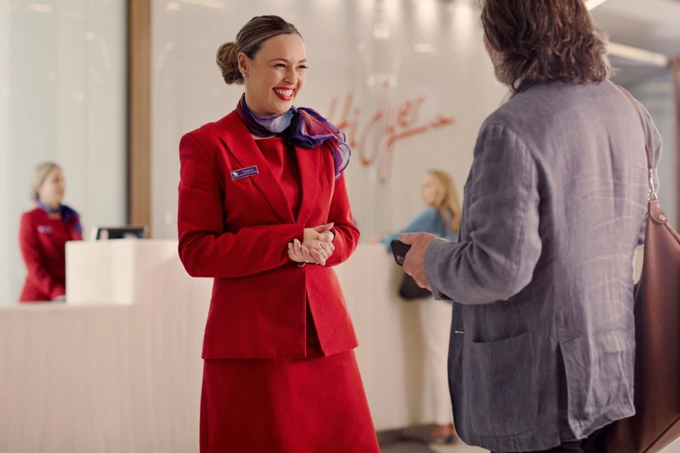 Smiling woman in red suit interacts with a man in a grey jacket at a modern airline check-in desk. Interior shot, bright light.