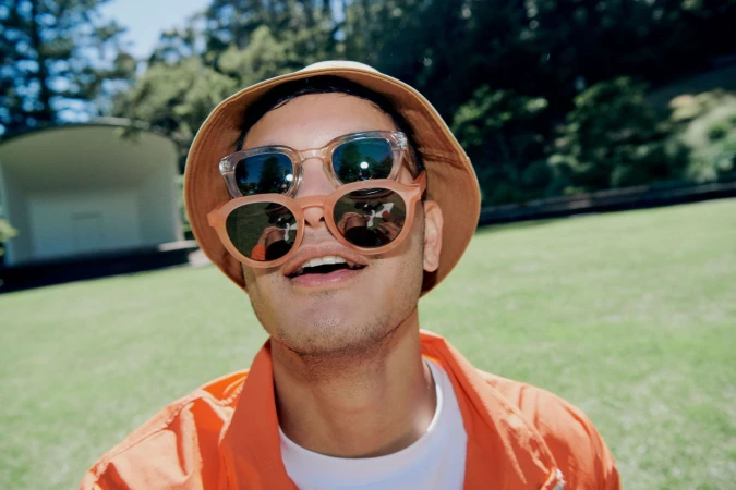 Close-up portrait of a man wearing two pairs of sunglasses, an orange bucket hat, and an orange jacket against a park lawn.