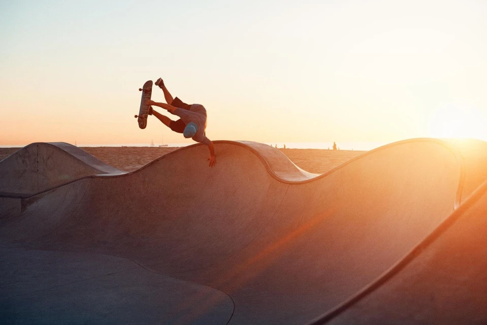 Adult male skateboarder performing a trick on a concrete ramp at sunset in a skatepark, wearing shorts and a light blue cap.