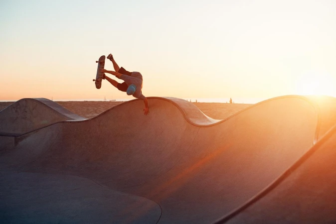 Adult male skateboarder performing a trick on a concrete ramp at sunset in a skatepark, wearing shorts and a light blue cap.