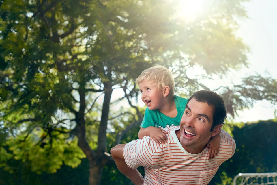 A cheerful father gives his blond-haired son a piggyback ride in a lush backyard with sun shining through the trees; both smiling.