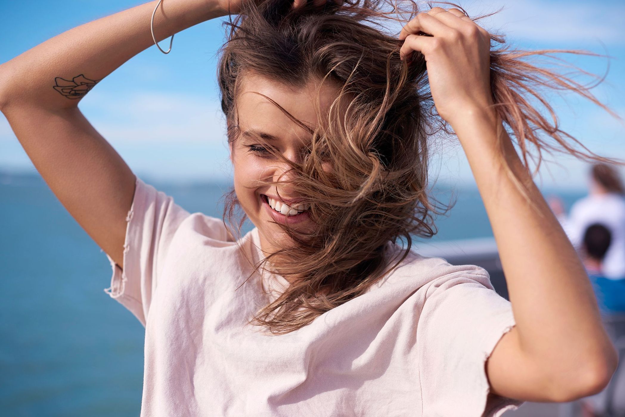 Smiling young woman with wavy brunette hair playfully touches her hair. Blue sky and ocean in the background during daytime.