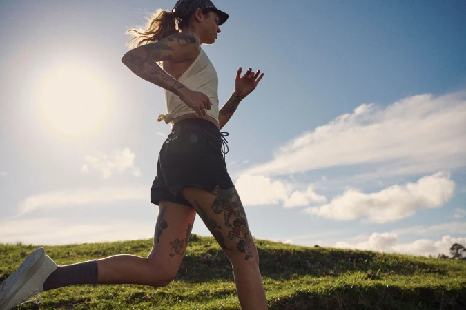 Tattooed woman running outdoors against a clear sky with sunflare, wearing a pale yellow top and dark shorts