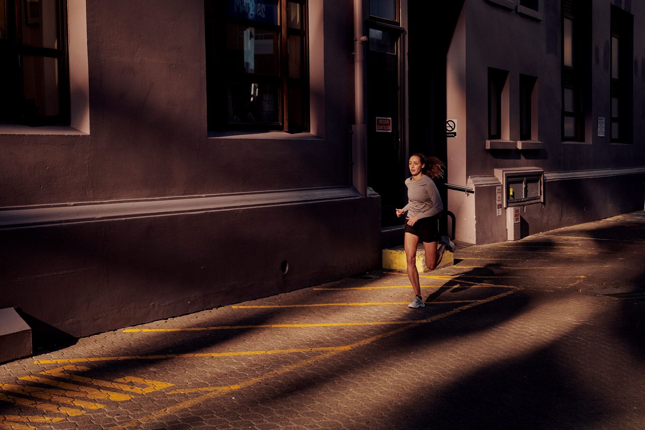 Athletic woman runs in urban alleyway with brick paving. Long shadows and golden light create contrast between the building walls.