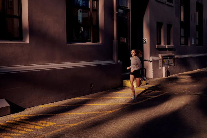 Athletic woman runs in urban alleyway with brick paving. Long shadows and golden light create contrast between the building walls.