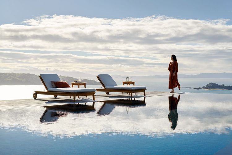Woman in red robe near infinity pool with lounge chairs overlooking ocean and island landscape under a cloudy sky on sunny day.