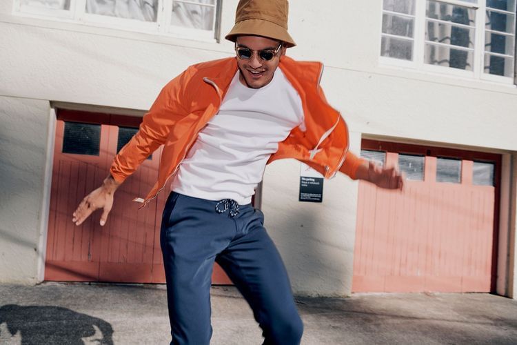 Man in orange jacket and bucket hat poses outside a pink door and white building, smiling in sunglasses in an urban environment.