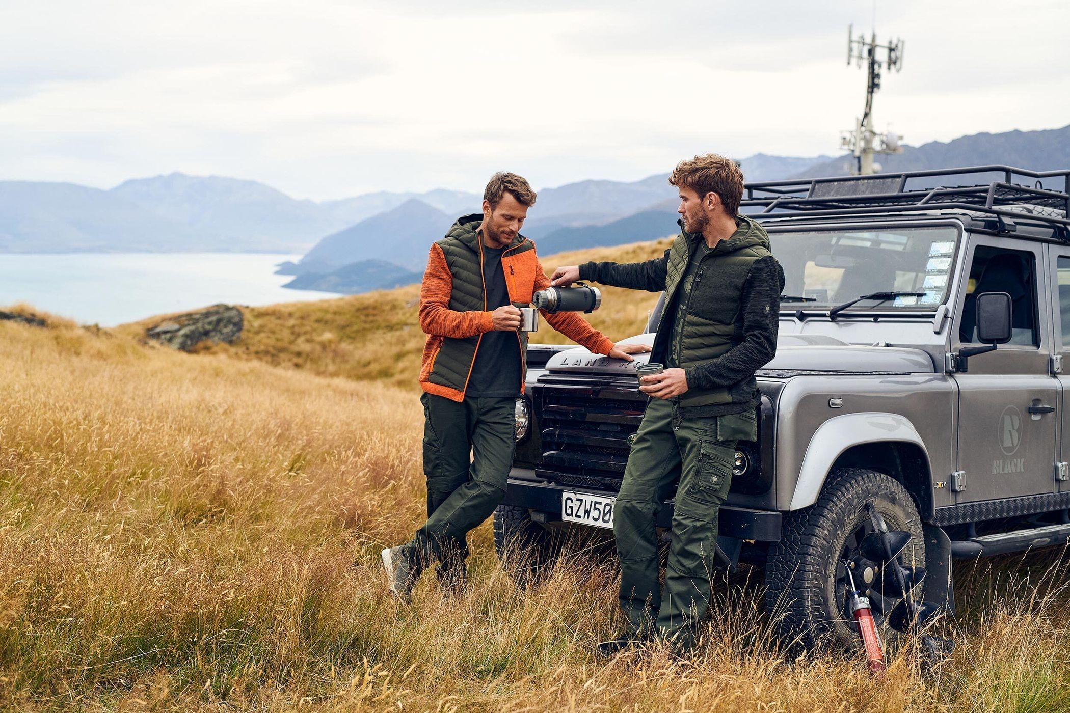 Two men in outdoor apparel next to a Land Rover in a mountain landscape, one pouring from a thermos with mountains and a lake behind them.