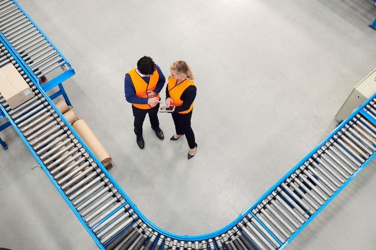 High-angle shot: two warehouse workers in safety vests review tablet near conveyor belts, in a bright industrial indoor setting.
