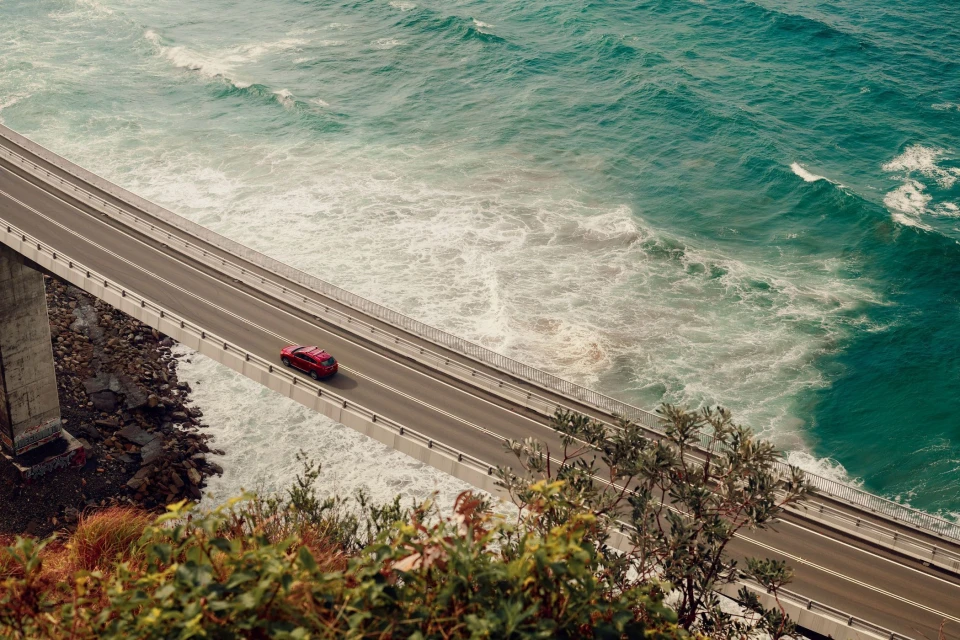 A red SUV drives along the Sea Cliff Bridge on a sunny day, offering picturesque views of the ocean and the New South Wales coastline.