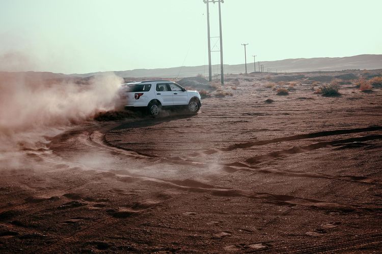 White SUV drives on a sand dune creating dust in a desert environment during daytime, with telephone poles in the background.