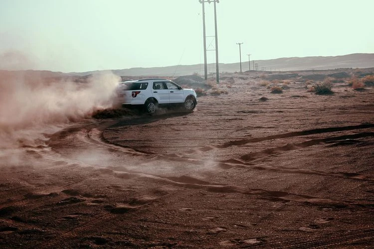 White SUV drives on a sand dune creating dust in a desert environment during daytime, with telephone poles in the background.
