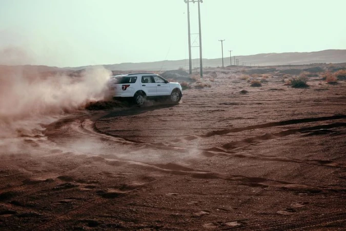 White SUV drives on a sand dune creating dust in a desert environment during daytime, with telephone poles in the background.