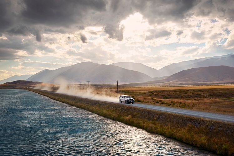 White SUV drives down rural dirt road with water canal beside it, mountain range and cloudy sky in the background, dust trailing behind.