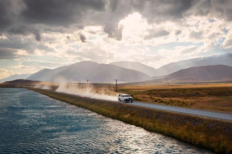 White SUV drives down rural dirt road with water canal beside it, mountain range and cloudy sky in the background, dust trailing behind.