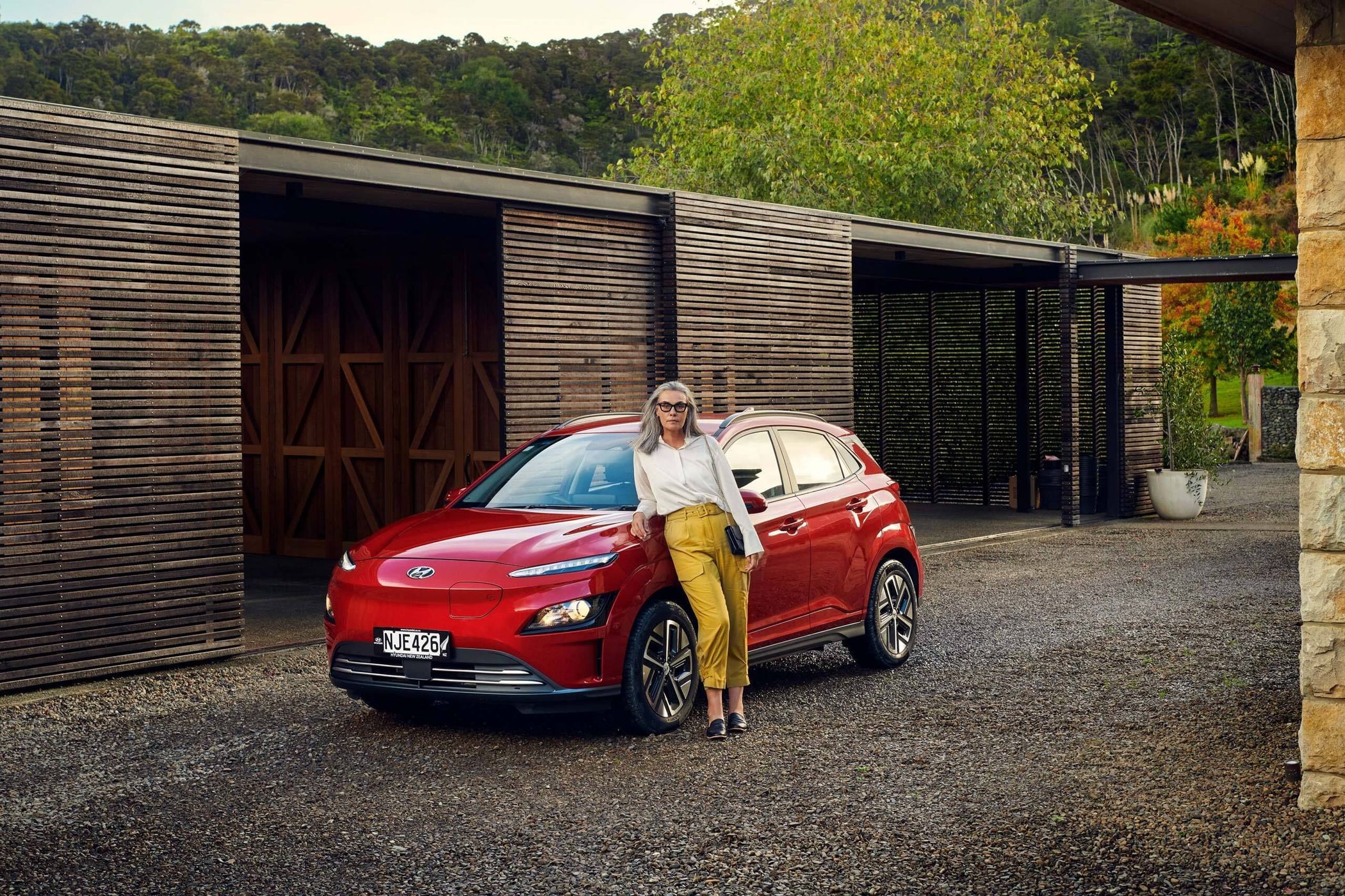 Red Hyundai Kona EV parked on a gravel driveway with an adult woman in yellow slacks leaning on it, against a modern wood building.