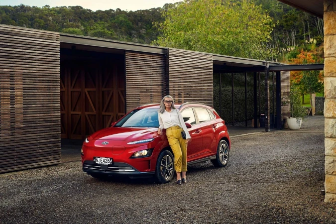 Red Hyundai Kona EV parked on a gravel driveway with an adult woman in yellow slacks leaning on it, against a modern wood building.
