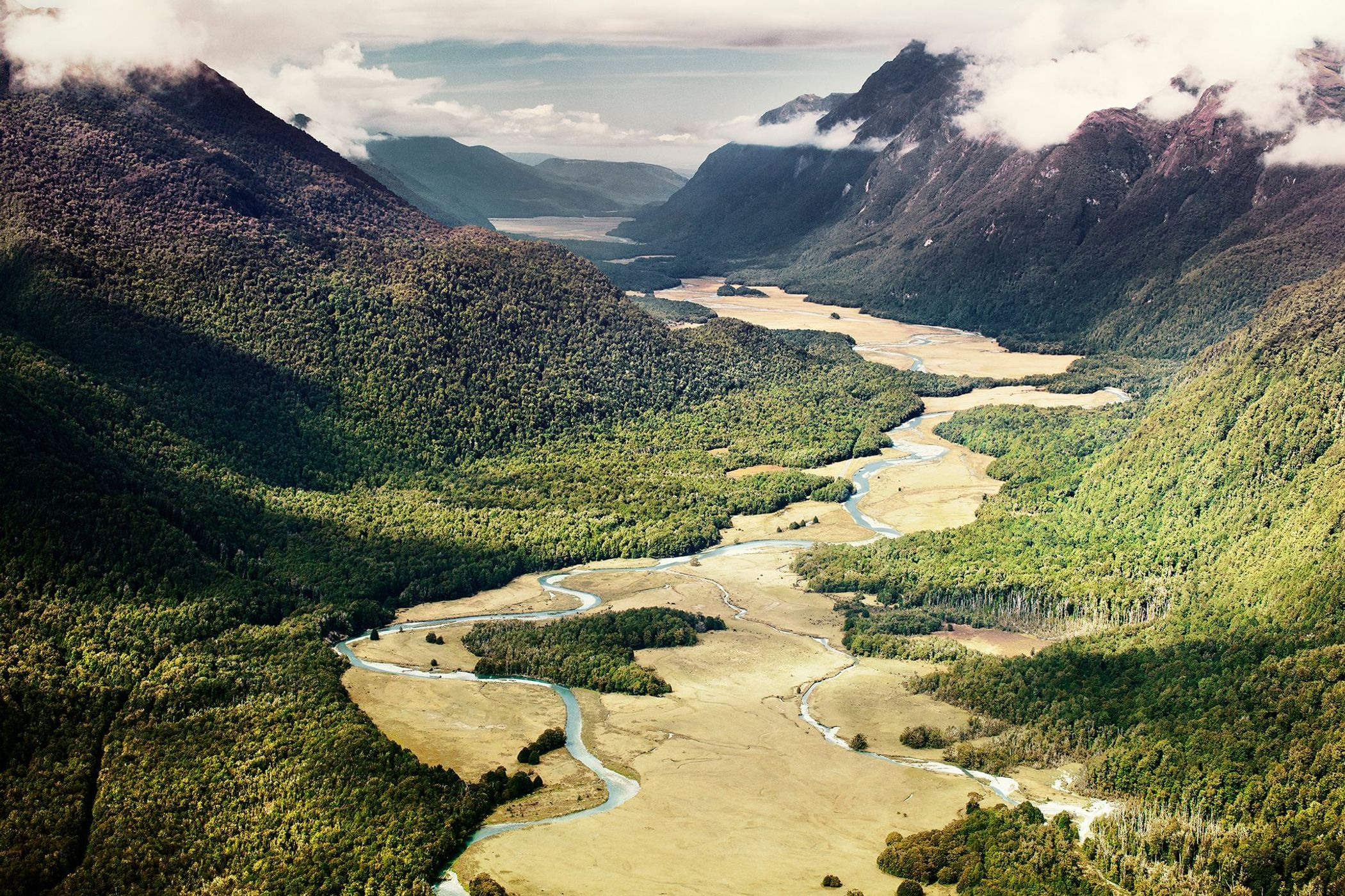 Aerial view of a river winding through a valley surrounded by lush green forests and mountains under a partly cloudy sky.