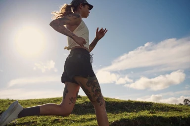 Tattooed woman running outdoors against a clear sky with sunflare, wearing a pale yellow top and dark shorts