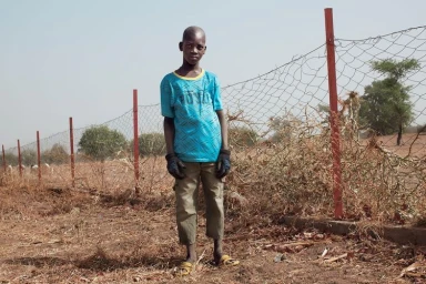 Young boy standing outdoors wearing a blue t-shirt and grey trousers against a wire fence and dry scrub landscape.
