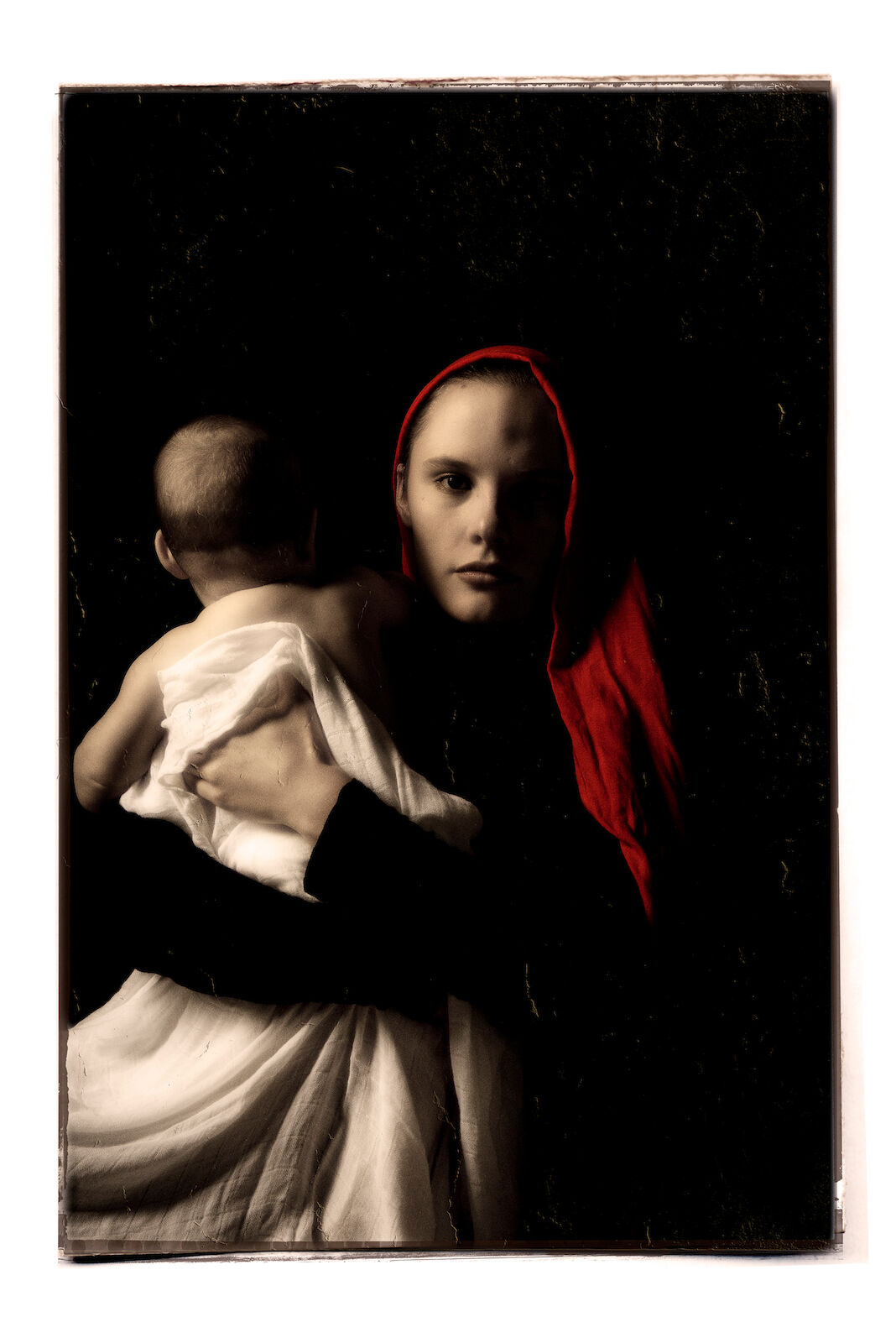 Woman holds a baby swaddled in white fabric against a dark background, with her face visible and a red headscarf accentuating the scene.