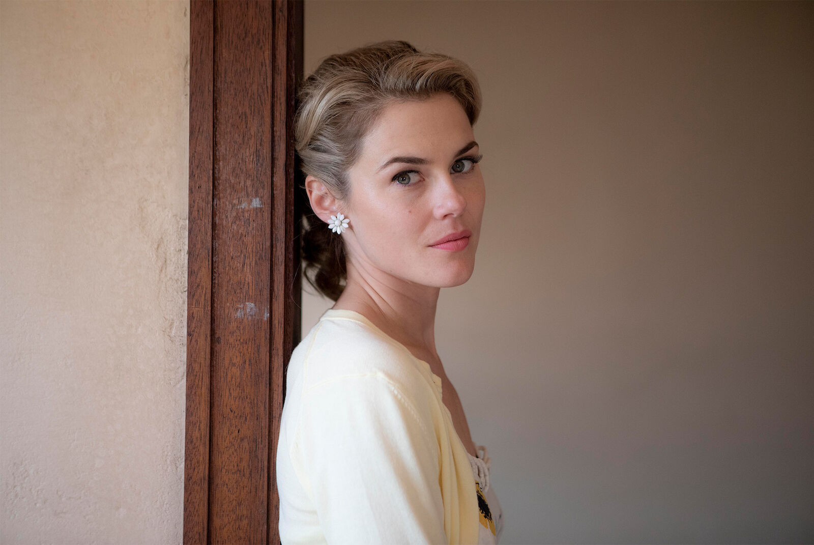 Portrait of an adult woman with blonde hair and white flower earrings, wearing a yellow cardigan, standing indoors by a doorway.