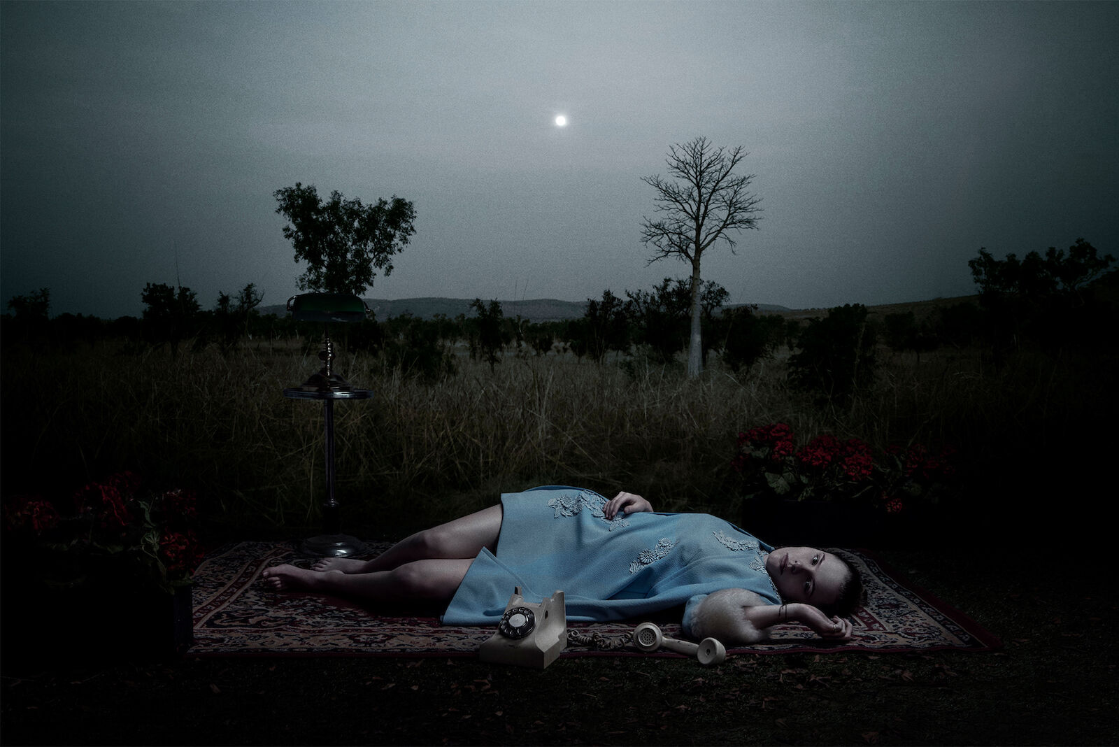 Woman lies on a rug with a vintage telephone in a field at night with a dark sky and trees in the distance under moonlight.