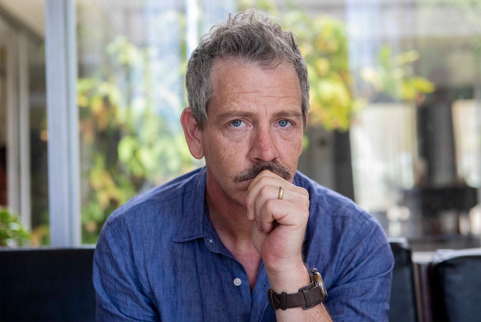 Portrait of a mature man with graying hair and a mustache, wearing a blue shirt, posing indoors with natural light and foliage in background.