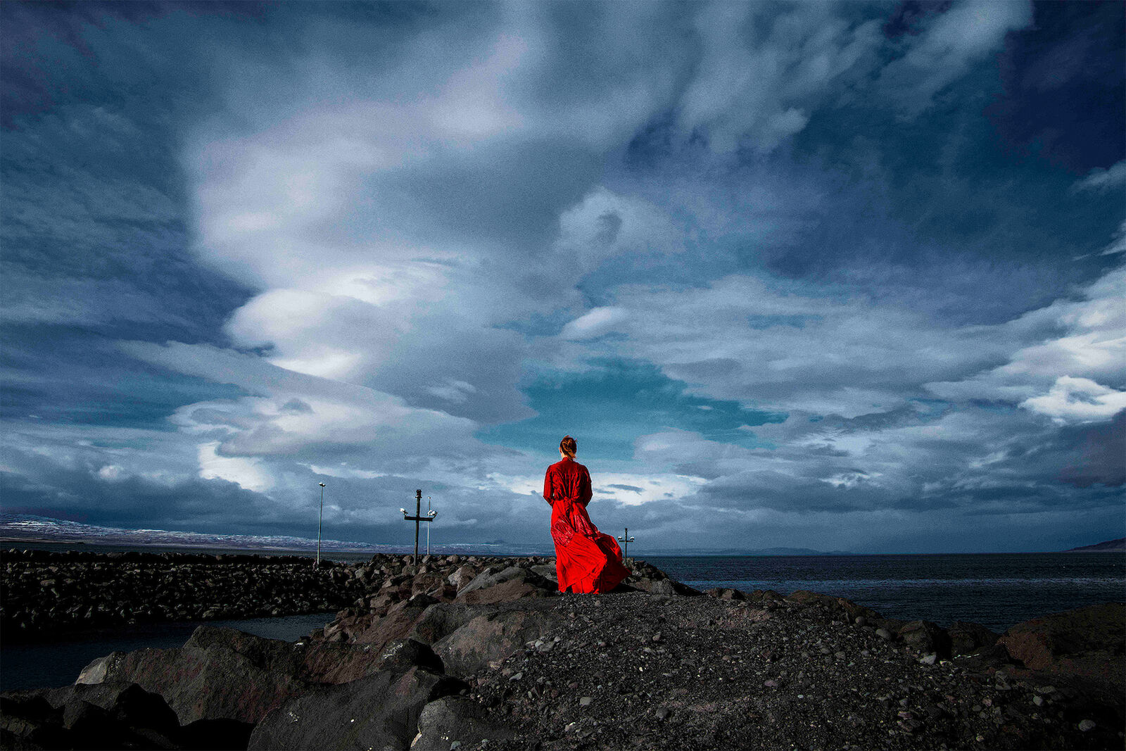 Woman in flowing red dress standing on rocky coast under dramatic cloudy sky, looking out to sea on an overcast day.