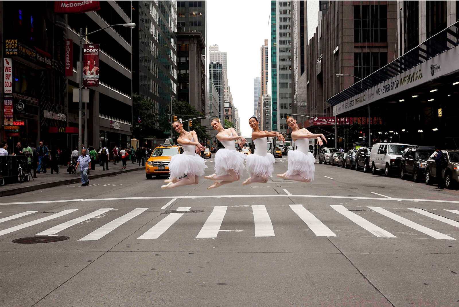 Four ballerinas in white tutus leap across a crosswalk in a bustling urban city street with tall buildings and yellow taxis.