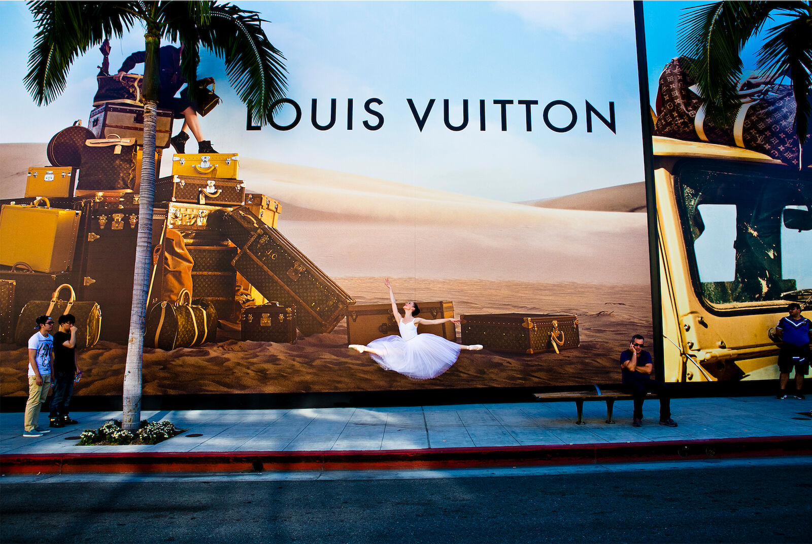 Ballerina leaps in front of a Louis Vuitton billboard featuring vintage luggage and a desert scene, with people on the sidewalk.