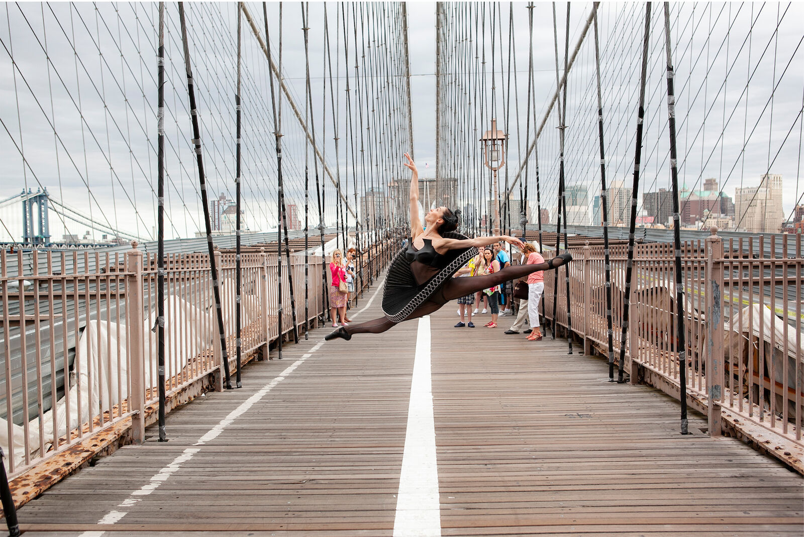 A woman in a black leotard leaps in the air on the Brooklyn Bridge, New York City, with the city skyline visible in the distance.