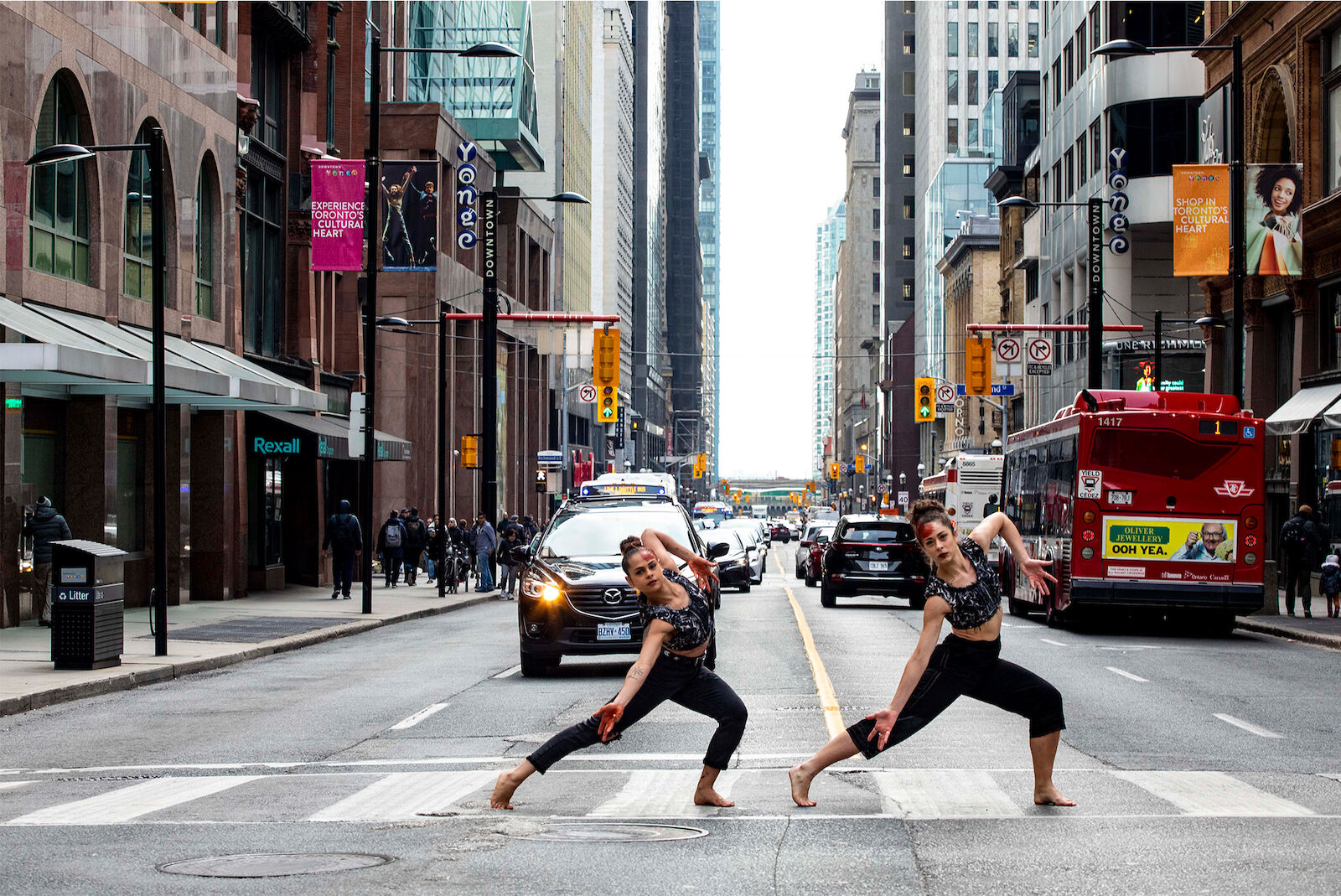 Two barefoot dancers perform on crosswalk in urban setting. Cars and a bus are on the street behind them with tall buildings in background.
