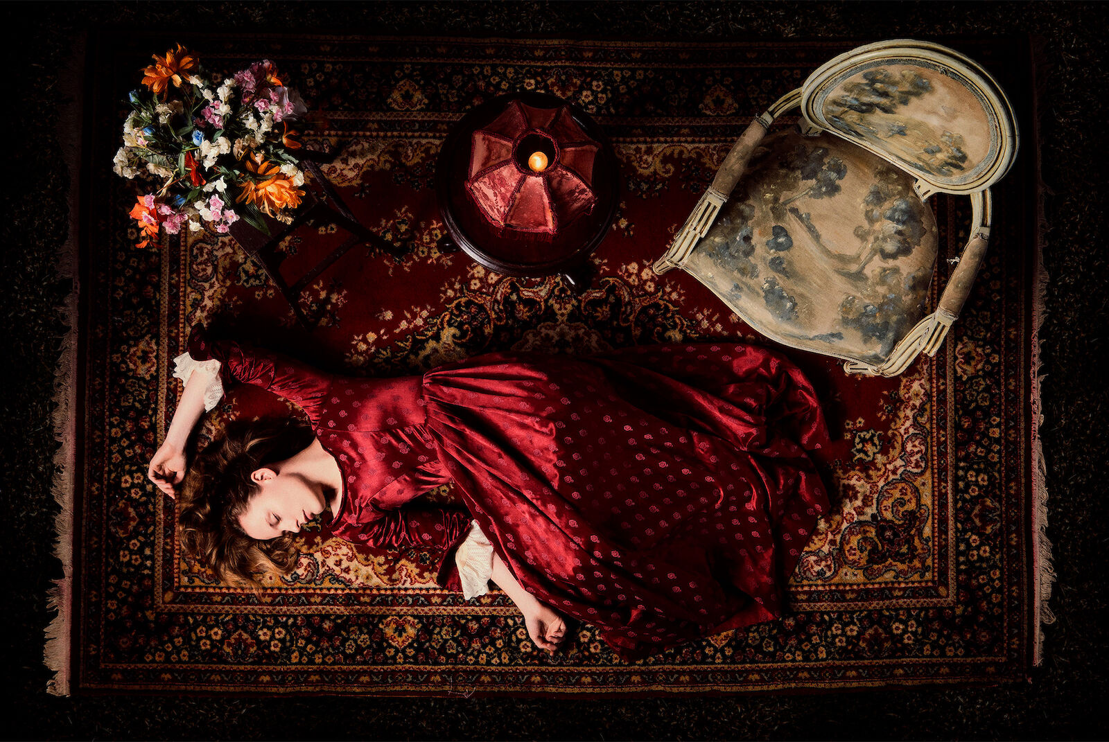 Overhead shot of a woman in a red dress lying on a patterned rug, with flowers, a candle, and an antique chair in a dark setting.