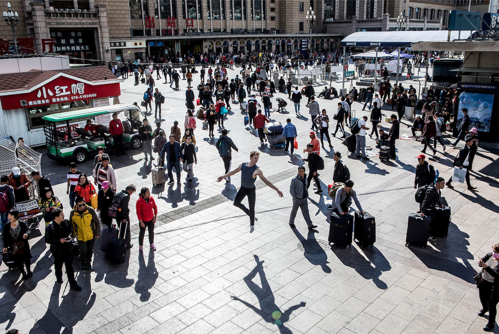 Wide shot of an outdoor plaza with a large group of pedestrians, including a male dancer jumping mid-air. Suitcases are visible.