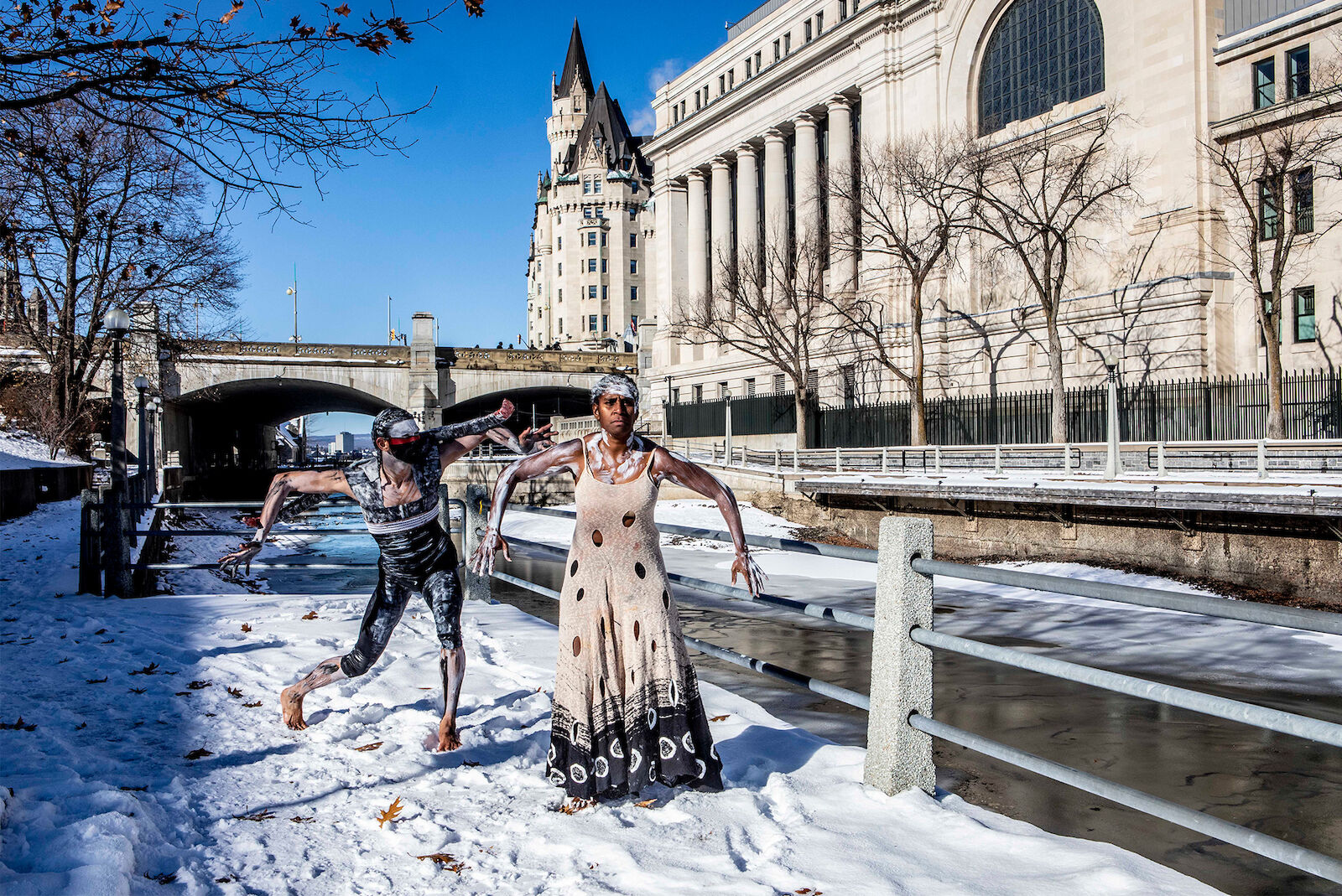 Two performers pose in wintery urban scene with stone buildings and snow, one in black and white body paint, one in a light dress.