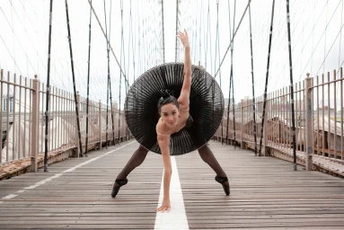 Ballet dancer in a large black tutu posing on a wooden pedestrian bridge walkway with suspension cables.