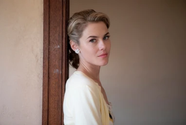 Portrait of a woman wearing white floral earrings and a light-colored top leaning against a dark wooden doorframe.