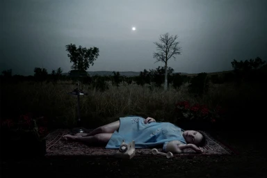 Woman in a blue dress lying on an ornate rug outdoors in a field at night with a rotary telephone and a glowing moon in the sky