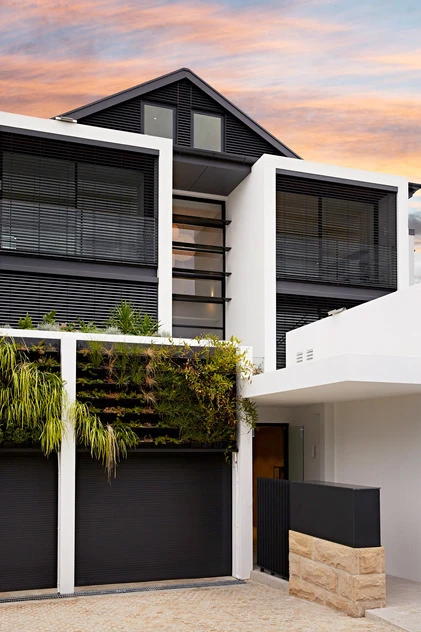 Modern house with white facade, black garage doors, and a green wall set against a colorful sunset sky. Architecture and design.