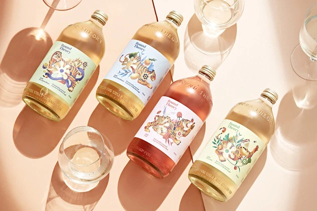 Four bottles of Bound Theory beverages with colorful label illustrations of tigers on a peach-colored surface with two drinking glasses.