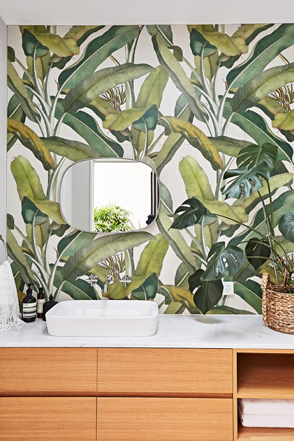 Bathroom interior featuring a white countertop, vessel sink, decorative mirror, wooden cabinets, green tropical-leaf wallpaper, and potted plants.