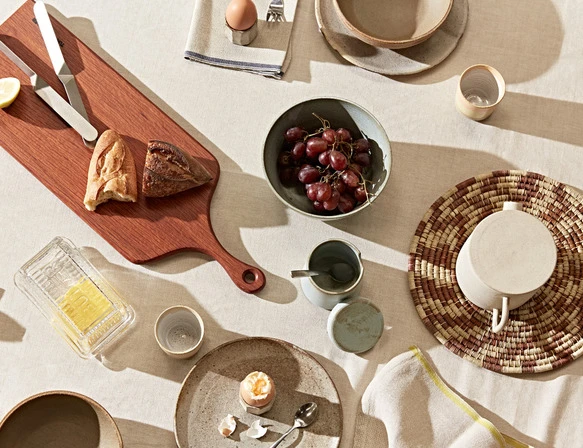 Overhead shot showcases breakfast still life with bread, fruit, boiled eggs, butter on wooden boards, plates, bowls and linen on table.
