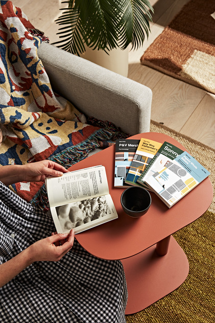 Person sitting on a couch reading an open book on a red side table with three books and a black bowl in an interior setting.