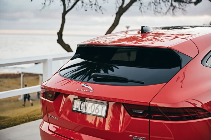 Red Jaguar E-Pace rear view by the coast with ocean backdrop and people walking in background. Tailgate, license plate and logo visible.
