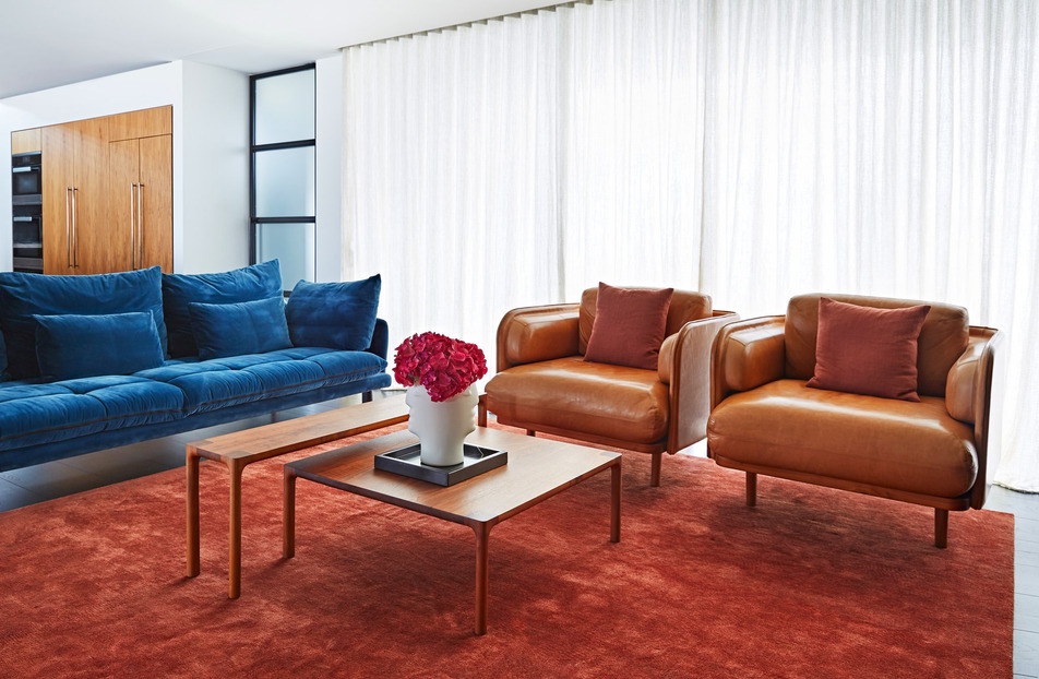 Living room interior showcasing a blue sofa, two brown leather armchairs with pillows, wooden coffee tables, and a red rug.