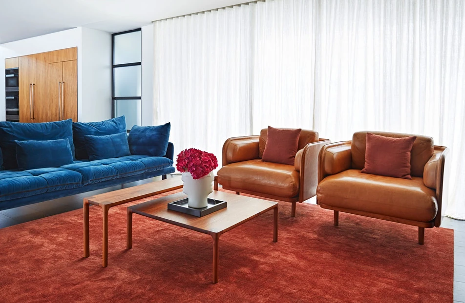 Living room interior showcasing a blue sofa, two brown leather armchairs with pillows, wooden coffee tables, and a red rug.