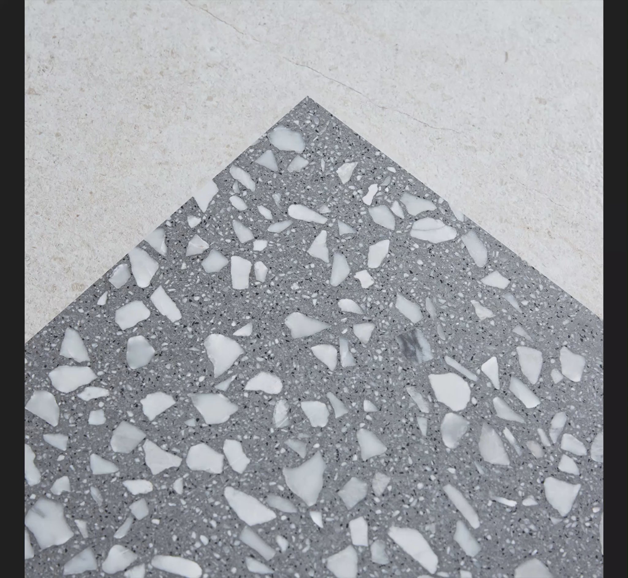 Close-up of a gray terrazzo tile with large white marble chips next to a beige textured tile, showing contrast and material detail.