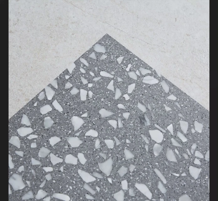Close-up of a gray terrazzo tile with large white marble chips next to a beige textured tile, showing contrast and material detail.