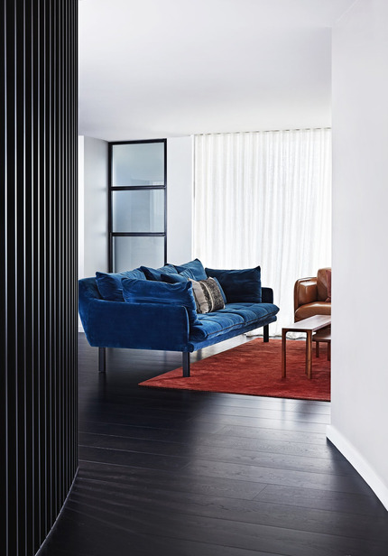 Living room interior with a blue velvet sofa, dark wood floors, a rust-colored rug, and white walls, bathed in soft, natural light.